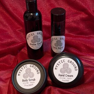 May include: A selection of Mystic Ground skincare items, comprising a gel cleanser, skin repair, body scrub, and hand cream. Each product is in a black container with a white label, displaying the brand name and product information. The items are arranged on red fabric.