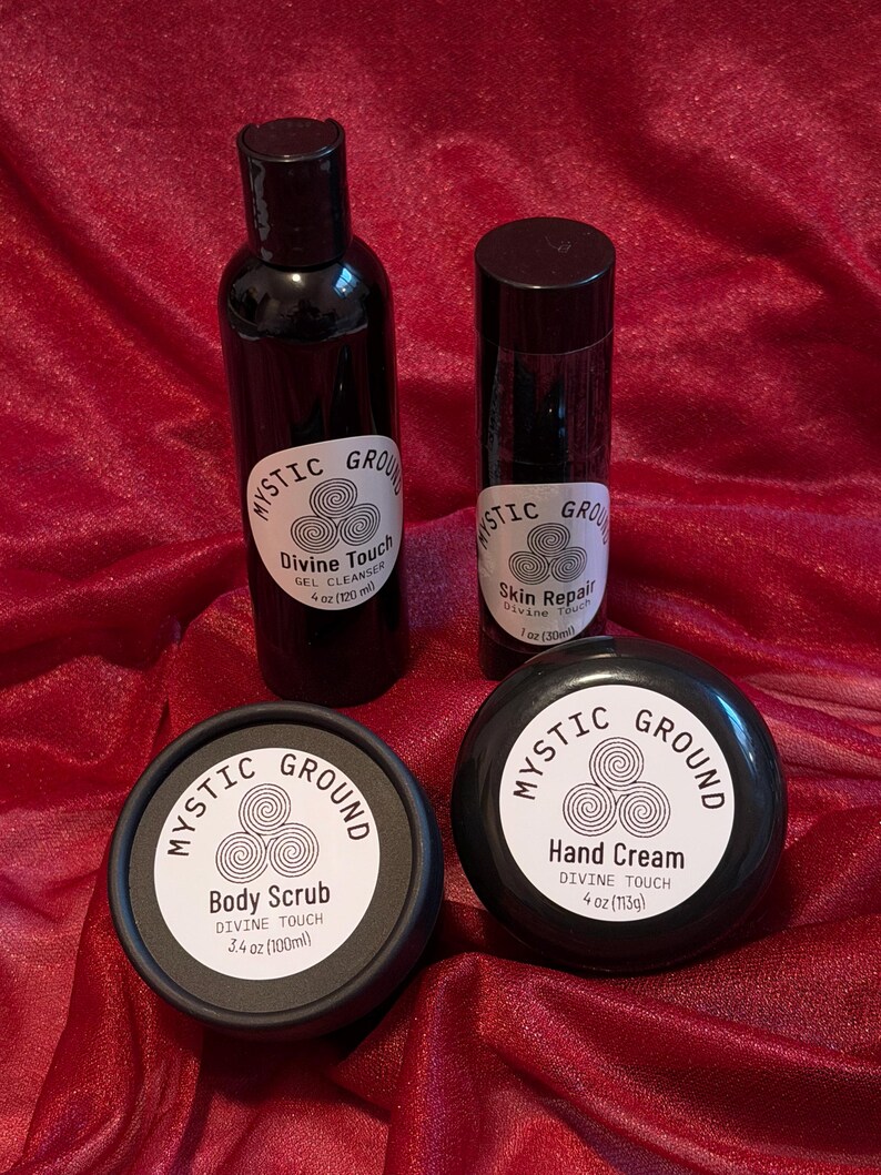May include: A selection of Mystic Ground skincare items, comprising a gel cleanser, skin repair, body scrub, and hand cream. Each product is in a black container with a white label, displaying the brand name and product information. The items are arranged on red fabric.