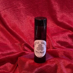 May include: A black cylindrical bottle with a white label that reads "Mystic Ground Skin Repair Divine Touch" and "1 oz (30ml)". The bottle is set against a red, textured fabric background.