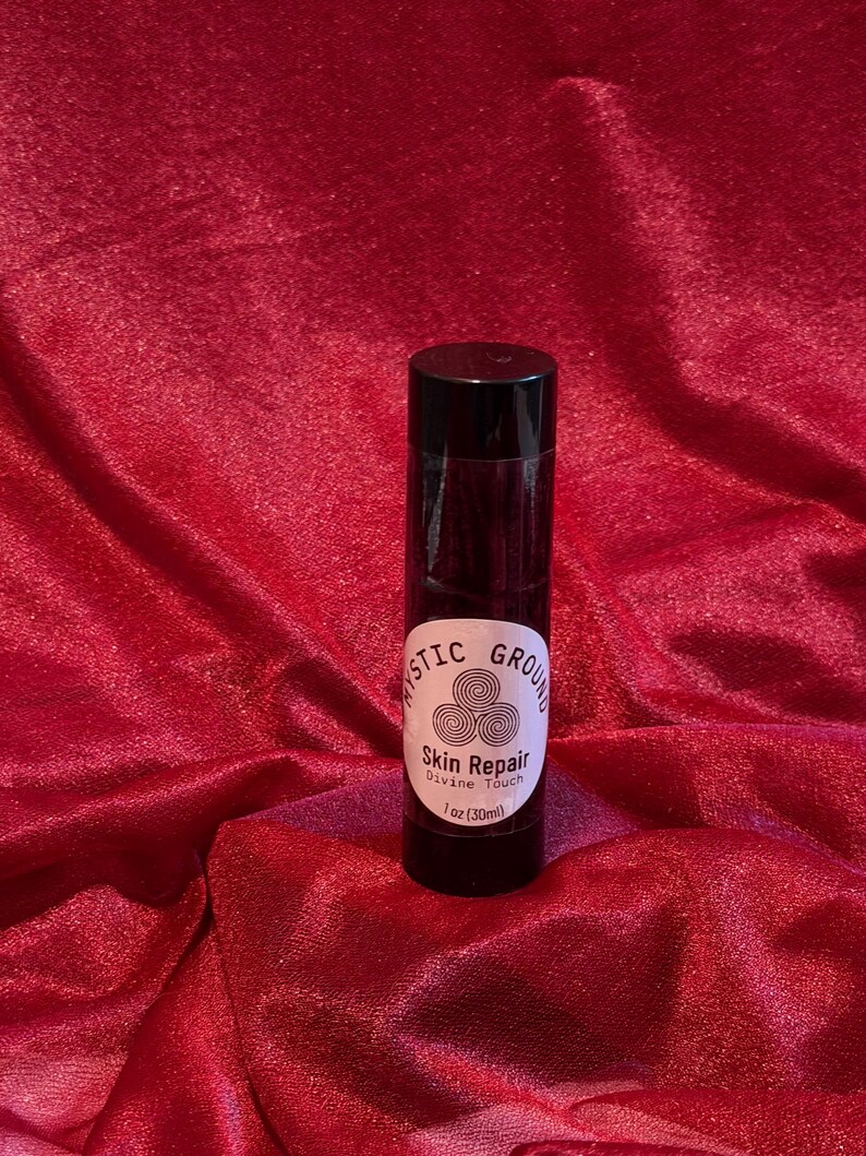 May include: A black cylindrical bottle with a white label that reads "Mystic Ground Skin Repair Divine Touch" and "1 oz (30ml)". The bottle is set against a red, textured fabric background.