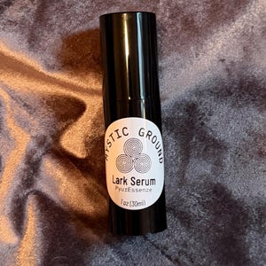 May include: A black serum bottle with a white label. The label reads "MYSTIC GROUND" above "Lark Serum" and "PyuzEssenze". The bottle contains 1 fl oz (30ml) of serum. The background is a textured, dark purple fabric.