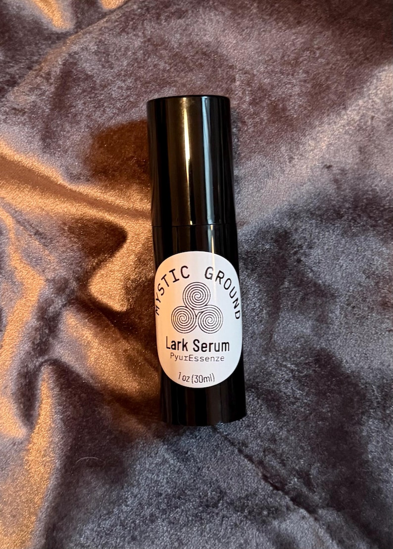 May include: A black serum bottle with a white label. The label reads "MYSTIC GROUND" above "Lark Serum" and "PyuzEssenze". The bottle contains 1 fl oz (30ml) of serum. The background is a textured, dark purple fabric.