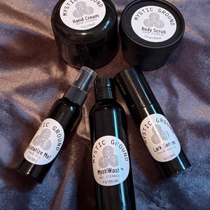 May include: An assortment of black cosmetic products from Mystic Ground, including hand cream, body scrub, face oil, gel cleanse, and lark serum. Each product has a white label with the brand name and product details. The products are arranged on a textured, dark purple surface.