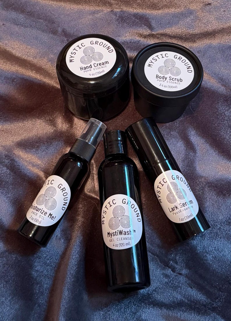 May include: An assortment of black cosmetic products from Mystic Ground, including hand cream, body scrub, face oil, gel cleanse, and lark serum. Each product has a white label with the brand name and product details. The products are arranged on a textured, dark purple surface.