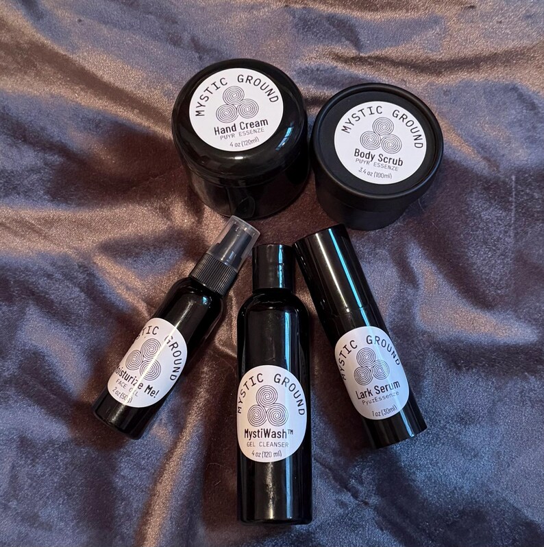 May include: An assortment of black skincare products from Mystic Ground, including hand cream, body scrub, a face mist, a gel cleanser, and a serum. Each product has a white label with the brand name and product details. The products are arranged on a dark, textured surface.