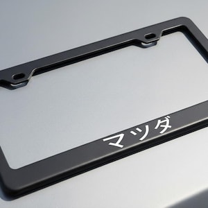 May include: Black license plate frame with the Japanese characters for Mazda in white. The frame is rectangular with rounded corners and mounting holes at the top.
