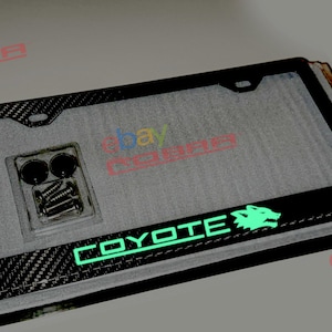 Peut inclure: Cadre de plaque d'immatriculation en fibre de carbone noire avec un logo "COYOTE" vert et une silhouette de loup. Le cadre est livré avec des vis et des capuchons.