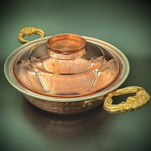 May include: A copper pot with a lid and two ornate handles. The pot has a hammered finish and a shiny, reflective surface.