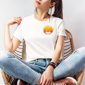 Pode incluir: T-shirt branco com um gráfico colorido de um sol, uma montanha e um animal. A camisa é de manga curta e tem gola redonda. A pessoa está usando jeans azuis e sapatos marrons.