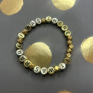 Peut inclure: Un bracelet en perles dorées et argentées avec les mots "I Was There" écrits dessus.