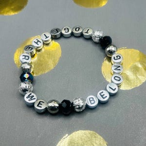 Op de afbeelding: Een zilveren kralenarmband met de woorden "WE BELONG TOGETHER" gespeld in individuele letterkralen. De armband bevat zilveren, zwarte en iriserende kralen. De armband ligt op een grijze ondergrond met gouden accenten.