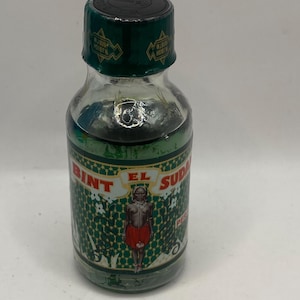 Puede incluir: Una pequeña botella de vidrio transparente con tapa y etiqueta verde oscuro. La etiqueta presenta el texto "BINT EL SUDAN" y una ilustración de una persona. La botella tiene un diseño verde y blanco.