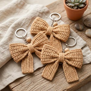 May include: Three tan crochet bow keychains, each featuring a textured yarn bow, a single round wooden bead, a silver-toned metal chain, and a circular split ring. The bows are crafted with a consistent stitch pattern and have two hanging tails.