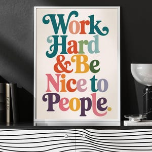 May include: A framed print with the words "Work Hard & Be Nice to People" in colorful, retro-style lettering. The artwork is displayed on a black and white striped cabinet, with books and a modern lamp nearby.
