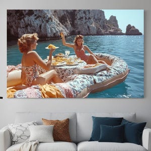 Op de afbeelding: Twee vrouwen in badpakken genieten van een maaltijd met pasta op een drijvend vlot in een turquoise blauwe zee. Het vlot is versierd met bloemenpatronen en heeft een witte tafel met borden pasta en een glas wijn.