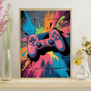 May include: A framed art print featuring a pink and blue video game controller against a colorful graffiti-style background. The controller has blue buttons and a black outline. The background has splashes of orange, blue, green, and pink.