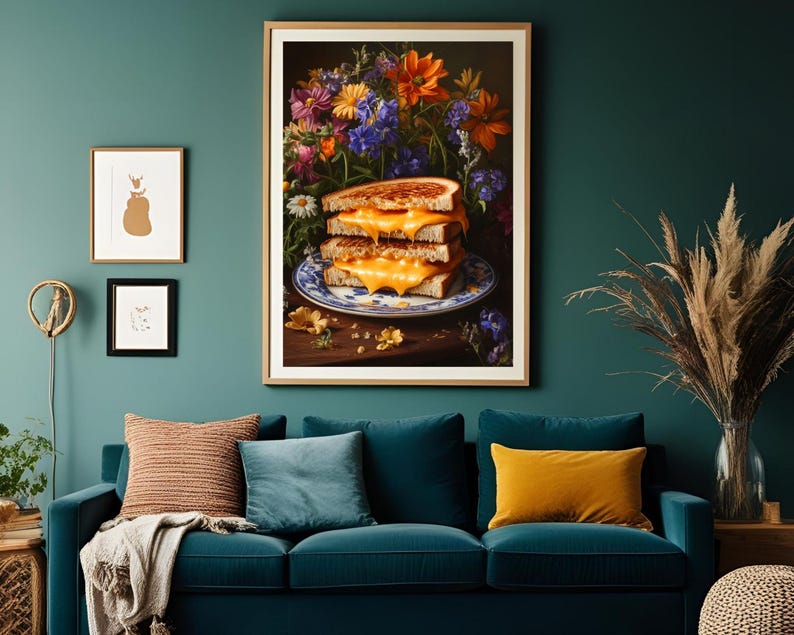 K&ouml;nnte beinhalten: Ein gerahmter Kunstdruck zeigt einen Stapel von drei gegrillten K&auml;sesandwiches auf einem blau-wei&szlig;en Teller, vor einem Hintergrund aus bunten Blumen. Das Kunstwerk h&auml;ngt &uuml;ber einem teal-farbenen Sofa mit Kissen und schafft eine gem&uuml;tliche Wohnzimmerszene.