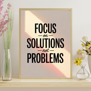 Może przedstawiać: Oprawiony wydruk z napisem "FOCUS on SOLUTIONS not PROBLEMS" czarnymi literami. Rama jest złota. Wydruk znajduje się na drewnianej półce z wazonami z kwiatami po obu stronach.