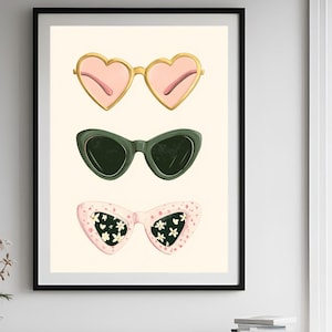 Puede incluir: Una impresión artística enmarcada con tres pares de gafas de sol. El par superior tiene monturas doradas en forma de corazón y lentes rosas. El par del medio es de estilo ojo de gato verde oscuro. El par inferior es rosa con detalles florales blancos.