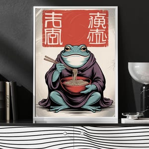 May include: A framed art print featuring a blue frog in a purple robe, holding chopsticks and a red bowl of noodles. The artwork has Japanese text in a red square at the top. The frog is the central focus of the illustration.