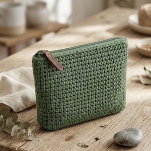 May include: A sage green rectangular zippered pouch featuring a textured crochet knit pattern. The pouch includes a matching green zipper closure and a small brown leather pull tab attached to the metal zipper slider for easy opening and closing.