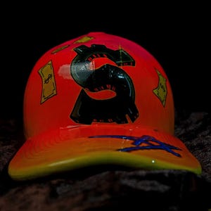 Puede incluir: Una gorra de béisbol roja y amarilla con un gráfico de signo de dólar negro. La gorra tiene una visera curva y una parte inferior amarilla.