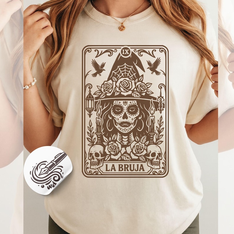 May include: Beige t-shirt featuring a brown graphic of a witch with a sugar skull face, roses, and the text "LA BRUJA". The design includes a witch hat, candles, and two skulls. The shirt has a round neck.