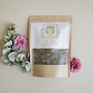 May include: A brown paper bag of Nora Organic Pregnancy Tea with a clear window, featuring a label with a cartoon illustration and the text "Tea Time With Nora". The bag is surrounded by pink flowers and green foliage.