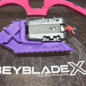 May include: A purple Beyblade X launcher with a black Beyblade on top. The launcher has a white "Beyblade X" logo and the text "Beyblade X" is printed in white on a gray surface.