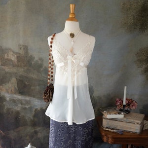 Op de afbeelding: Een witte kanten top met een satijnen strik en een rok met bloemenprint op een houten mannequin. De mannequin staat op een houten standaard met een rieten mand en een vogelkooi ernaast. Er staan ​​bruine laarzen op de vloer.