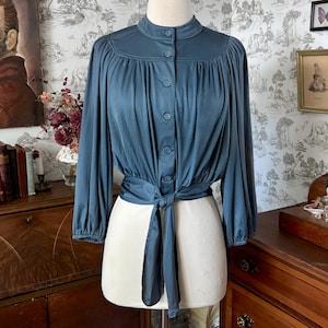 May include: A slate blue long-sleeve blouse featuring a high collar, a front button-down closure, and a tie-front waist detail. The garment has gathered fabric at the shoulders and cuffs, creating a soft, draped silhouette on a dress form.