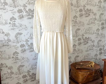 Könnte beinhalten: Ein cremefarbenes Vintage-Kleid auf einer Holzpuppe. Das Kleid hat ein strukturiertes Oberteil, lange Spitzenärmel und einen fließenden Rock. Das Kleid wird vor einer Tapete mit Landschaftsdesign präsentiert.
