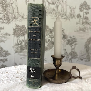 Puede incluir: Un libro antiguo titulado "The Poems of Longfellow" con una cubierta verde y letras doradas, junto a un candelero de latón que sostiene una vela blanca. El fondo presenta un papel pintado con dibujos.