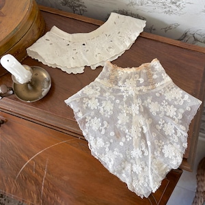 May include: Two antique white lace collars. One is a scalloped eyelet design, and the other is a square-shaped collar with a floral pattern. The collars are displayed on a dark wooden surface.