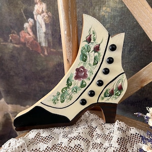 May include: A hand-painted wooden shoe with a black heel and toe. The shoe is decorated with a floral design in shades of pink, green, and white. There are black buttons on the side of the shoe.