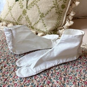 May include: A pair of white, split-toe tabi socks, made of a soft fabric, rest on a floral patterned quilt. The socks are designed to be worn with traditional Japanese footwear. A decorative pillow with green embroidery is in the background.