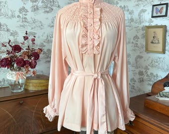 May include: A light pink blouse with long sleeves, a high ruffled collar, and a button-down front. The blouse features a cinched waist with a matching tie and is displayed on a wooden mannequin. The fabric has a slight sheen.