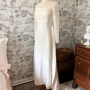 Puede incluir: Vestido de novia marfil vintage sobre un maniquí. El vestido presenta un corpiño de encaje con mangas largas y una falda fluida con cola. El vestido se exhibe frente a un fondo de pantalla.