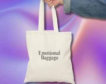 May include: A white tote bag with black text that reads "Emotional Baggage". The bag is being held by a hand against a purple and pink gradient background.