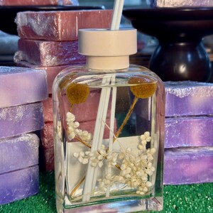 Puede incluir: Un difusor de vidrio transparente con tapa blanca y palitos blancos. La botella está llena de un líquido transparente y flores blancas. La botella está sobre una superficie verde.