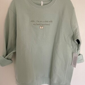 Peut inclure: Un sweat-shirt vert clair avec le texte "shhh...i'm on a date with my book boyfriend" et une petite illustration d'un livre.