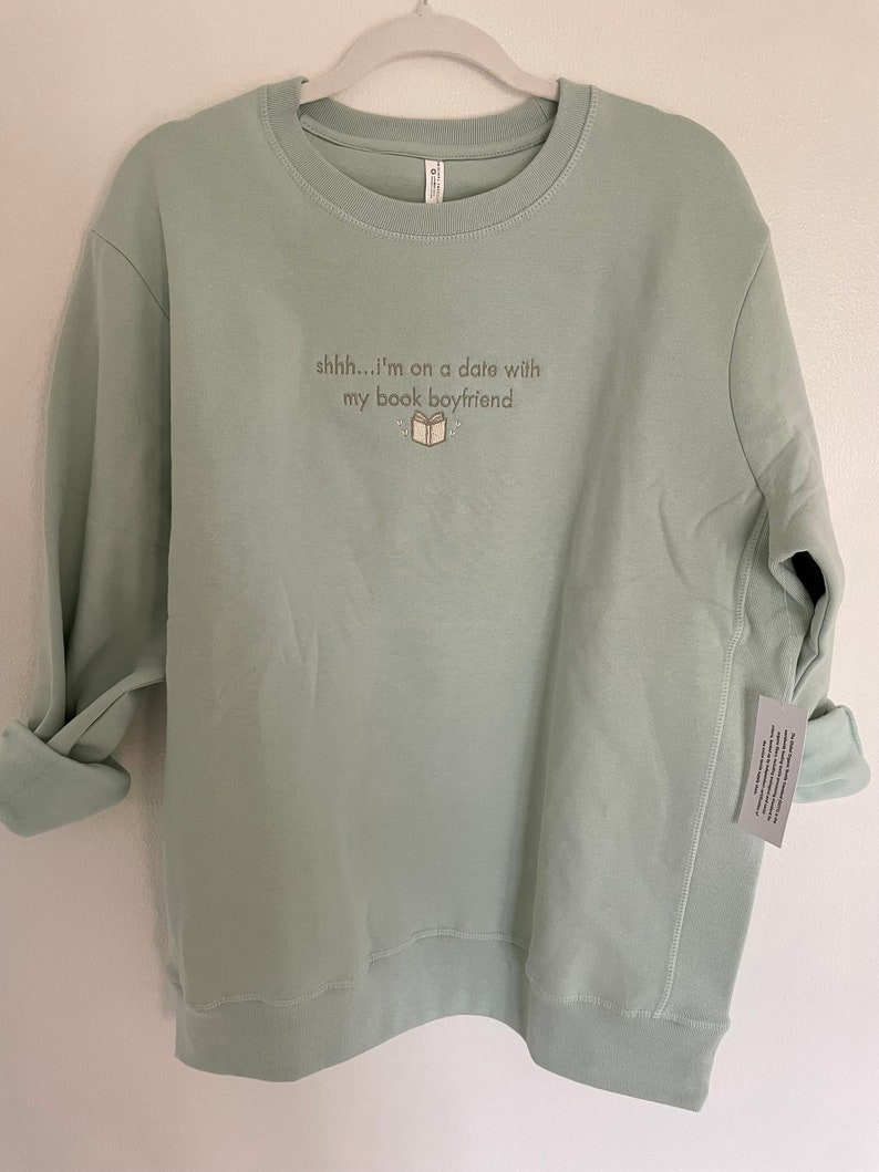 Peut inclure: Un sweat-shirt vert clair avec le texte "shhh...i'm on a date with my book boyfriend" et une petite illustration d'un livre.