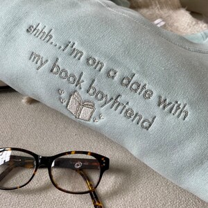 Peut inclure: Un sweat-shirt bleu clair avec le texte "shhh...i'm on a date with my book boyfriend" brod&eacute; en fil argent&eacute;. Une petite image brod&eacute;e d'un livre est &eacute;galement visible. Une paire de lunettes en &eacute;caille de tortue repose sur une surface beige.
