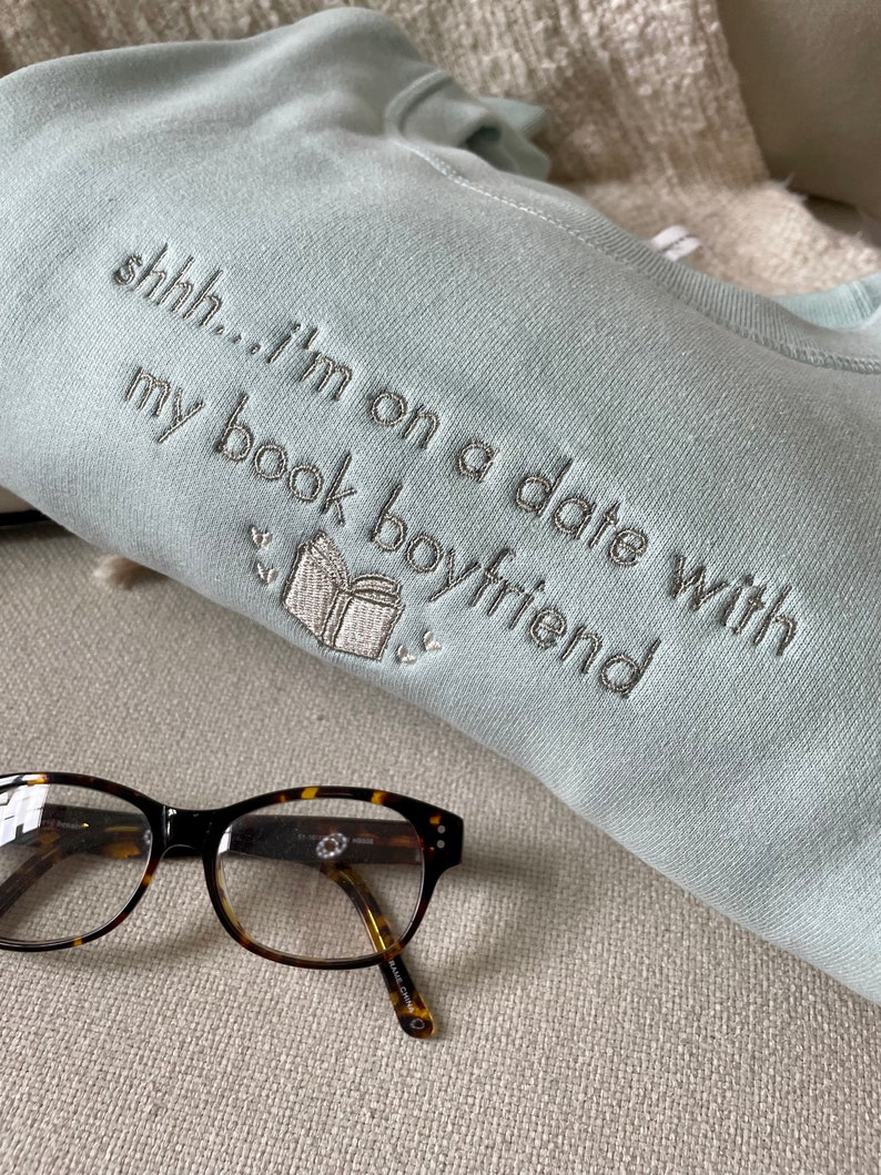 Peut inclure: Un sweat-shirt bleu clair avec le texte "shhh...i'm on a date with my book boyfriend" brod&eacute; en fil argent&eacute;. Une petite image brod&eacute;e d'un livre est &eacute;galement visible. Une paire de lunettes en &eacute;caille de tortue repose sur une surface beige.