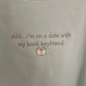 Peut inclure: Un t-shirt bleu clair avec le texte "shhh...i'm on a date with my book boyfriend" brod&eacute; sur le devant. Le texte est en fil brun et il y a une petite image brod&eacute;e d'un livre avec des c&oelig;urs au-dessus et en dessous.