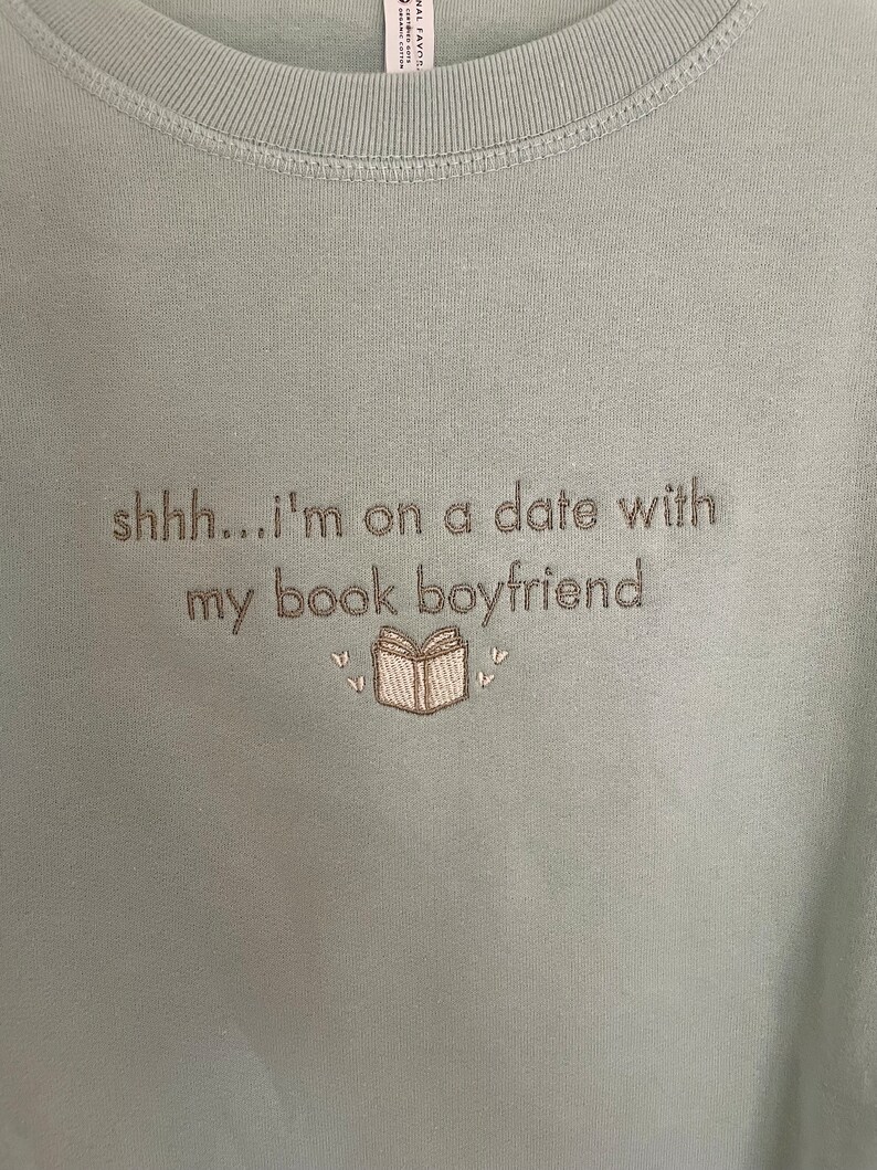 Peut inclure: Un t-shirt bleu clair avec le texte "shhh...i'm on a date with my book boyfriend" brod&eacute; sur le devant. Le texte est en fil brun et il y a une petite image brod&eacute;e d'un livre avec des c&oelig;urs au-dessus et en dessous.