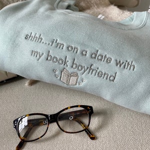 Peut inclure: Un sweat-shirt bleu clair avec le texte "shhh... i'm on a date with my book boyfriend" brod&eacute; sur le devant. Le sweat-shirt est pos&eacute; sur une surface brun clair avec une paire de lunettes en &eacute;caille de tortue.