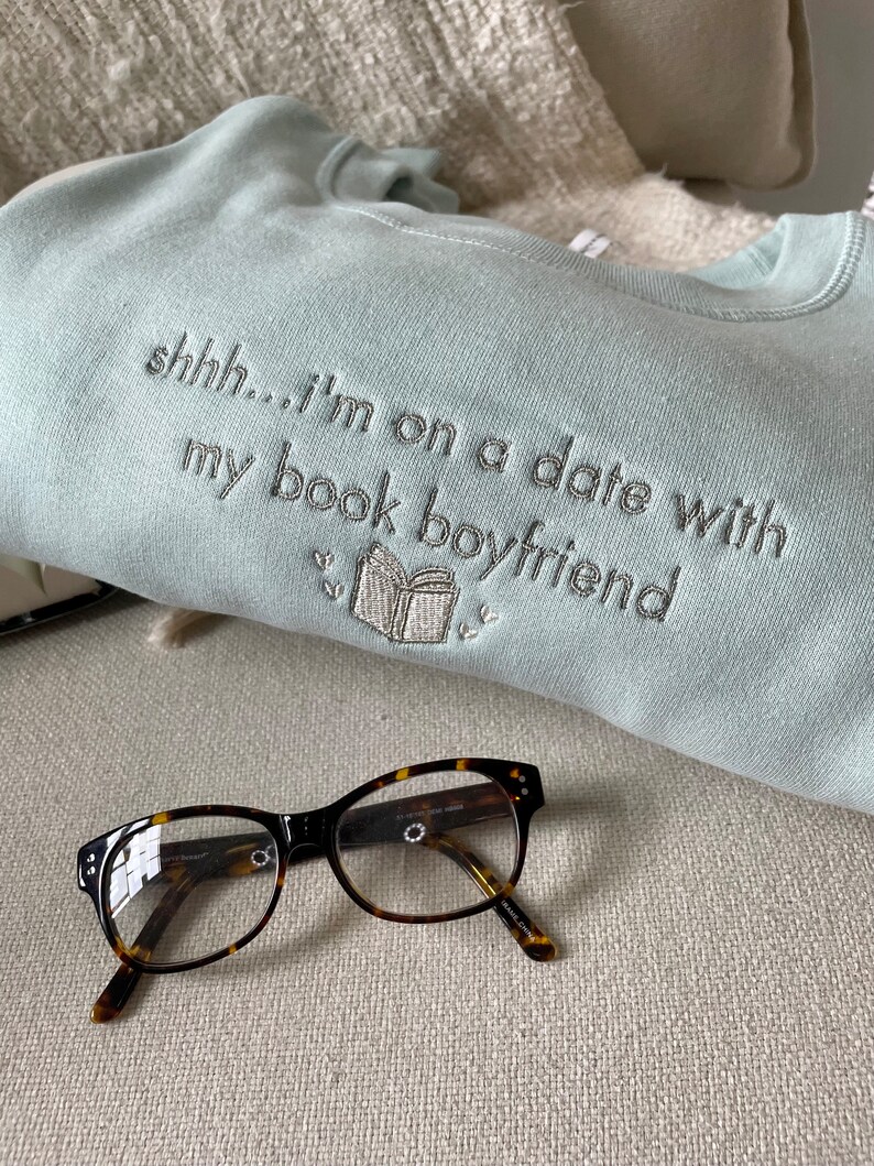 Peut inclure: Un sweat-shirt bleu clair avec le texte "shhh... i'm on a date with my book boyfriend" brod&eacute; sur le devant. Le sweat-shirt est pos&eacute; sur une surface brun clair avec une paire de lunettes en &eacute;caille de tortue.