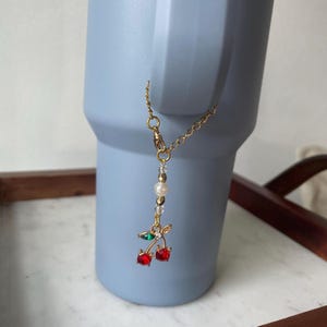 Könnte beinhalten: Eine goldene Kette mit einem Anhänger aus zwei roten Kirschen mit grünen Blättern und weißen Perlen. Die Kette ist an einem blauen Becher befestigt.