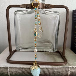 Puede incluir: Una botella de vidrio decorativa con un marco de metal marrón, con un dije de cuentas. El dije incluye cuentas turquesas, perlas y un colgante en forma de fresa azul claro con detalles dorados. La botella está vacía y transparente.